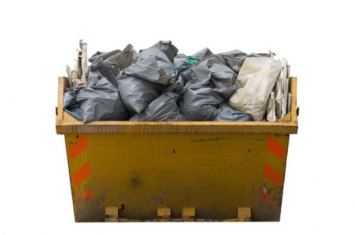 A large yellow skip bin, positioned on a flat surface, is filled with tightly packed black and grey refuse bags, along with some white plastic or fabric items visible among the waste. The skip shows signs of outdoor use, with minor dirt and wear on its exterior. The waste bags are piled towards the top edge of the bin, with some bags slightly bulging and others more compressed. The skip is situated outdoors, likely on a driveway or a waste collection area, with no background scenery visible. The image visually relates to rubbish removal services, such as those provided by Commercial Waste Ilford, serving the Ilford area. The environment and item arrangement emphasize the focus on waste disposal, fitting for a service advert or informational page regarding rubbish collection and recycling in the local area.