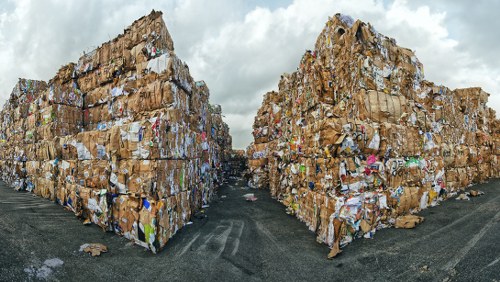 Two large stacks of compacted waste paper and cardboard, arranged in rectangular blocks on a paved outdoor surface. The stacks feature varied textures with visible corrugated cardboard, flat paper sheets, and crumpled material, predominantly in shades of brown, with some colourful paper pieces interspersed. The stacks are positioned on either side of a narrow pathway, with the sky overhead showing grey clouds, indicating an overcast day. The environment appears to be an industrial or waste processing yard, with the ground made of asphalt or concrete. This scene reflects the type of waste that Commercial Waste Ilford manages for recycling and disposal, supporting local rubbish removal needs around Ilford and the surrounding Essex area.