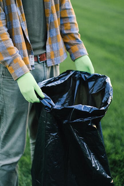 A person wearing a white glove and a blue wristwatch is holding a white plastic bag filled with garden waste, including small branches, green and brown leaves, and some gravel or soil. The bag is part