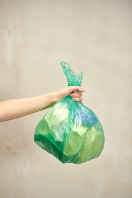 A person’s arm extending from the left side of the image holding a green plastic rubbish bag with blue patches, which appears to be filled and tied at the top with a twist tie, against a plain, ligh