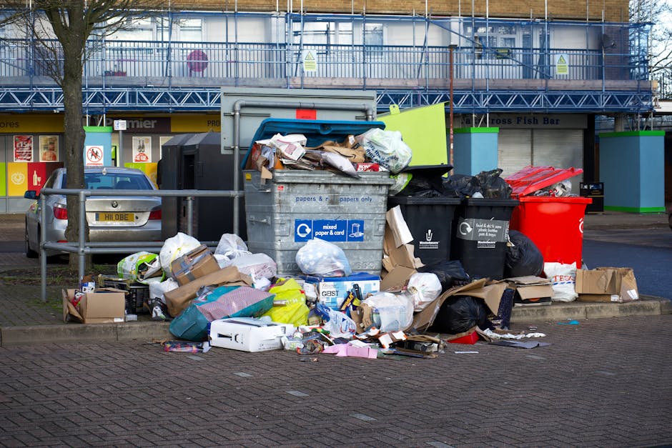 A middle-aged man with dark hair wearing a black T-shirt and dark trousers is seen using metal trash bins for rubbish disposal on a pavement. The man is leaning forward, reaching into one of the cylin